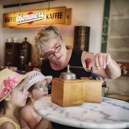 Zwei kleine Mädchen schauen zu, wie eine Frau mit einer Handmühle Kaffee mahlt.