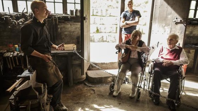 Zwei Personen mit Rollator und Rollstuhl hören in der Schmiede einerm Handwerker zu