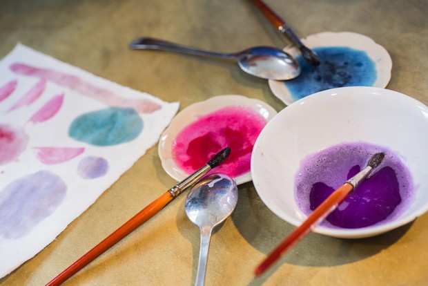 Auf einem Tisch stehen Schalen mit selbst hergestellte Farben, Pinsel und bemalte Papierbögen
