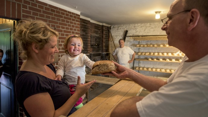 Eine Frau mit Kind auf dem Arm kauft in der Museumsbäckerei ein Brot. Der Bäcker reicht dem Kind das Brot über die Theke. Das Kind freut sich.