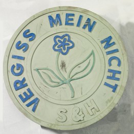 Ein rundes Firmenschild mit dem Spruch „Vergiss mein nicht“ und die Initialen „S & H“. In der Mitte ist ein stilisiertes Vergissmeinnicht.