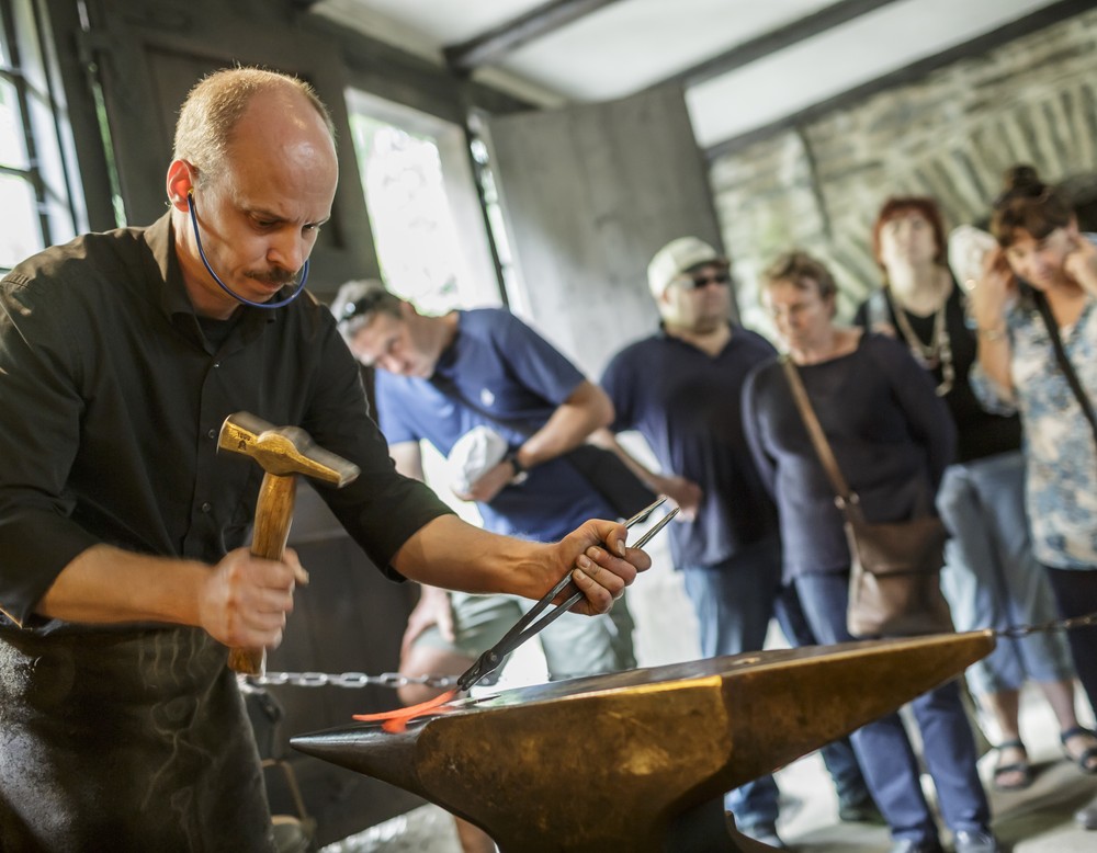 Ein Schmied mit Ohrenschutz führt einer Besuchergruppe mit Hammer und Zange auf einem Amboss sein Handwerk vor.