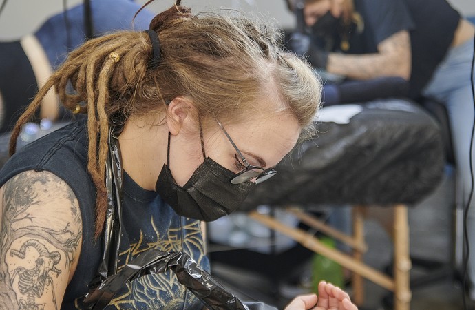 Eine Frau mit Dreadlocks, Brille und Mundschutz tätowiert einen Arm